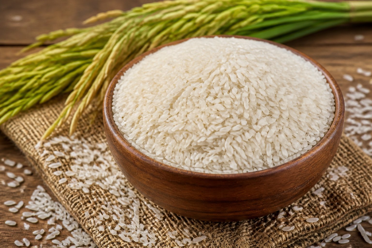 A wooden bowl filled with uncooked sona masoori rice grains on a jute fabric surface, with scattered rice grains and green rice stalks in the background.